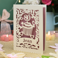 A personalised 80th birthday laser-cut greeting card for Jean, featuring a cream cupcake design with a pink background, displayed on a checked tablecloth with lit candles in the background.