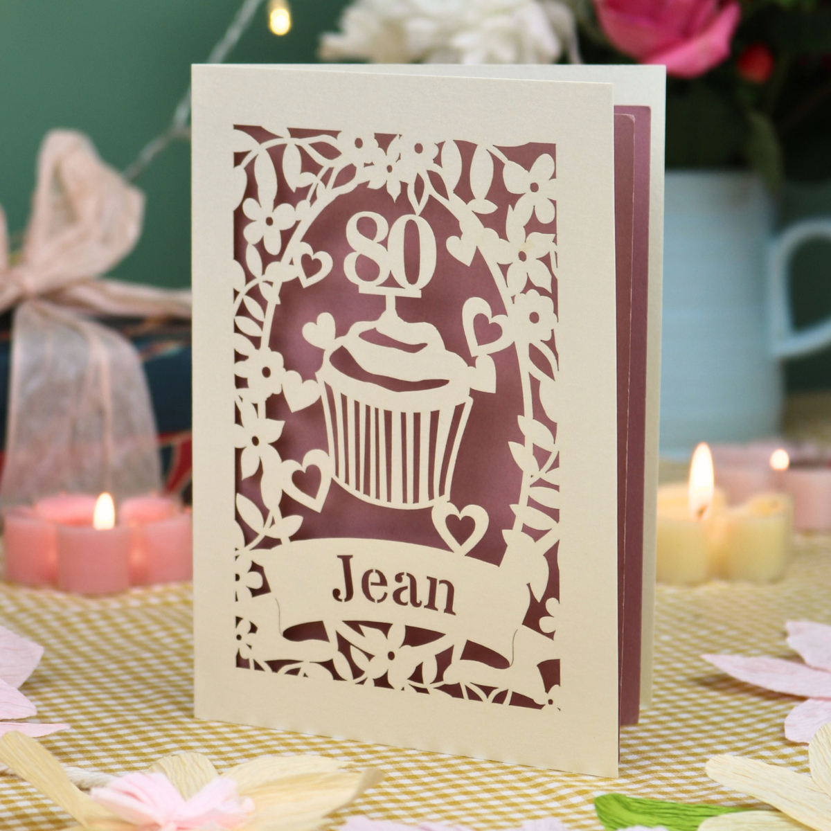 A personalised 80th birthday laser-cut greeting card for Jean, featuring a cream cupcake design with a pink background, displayed on a checked tablecloth with lit candles in the background.