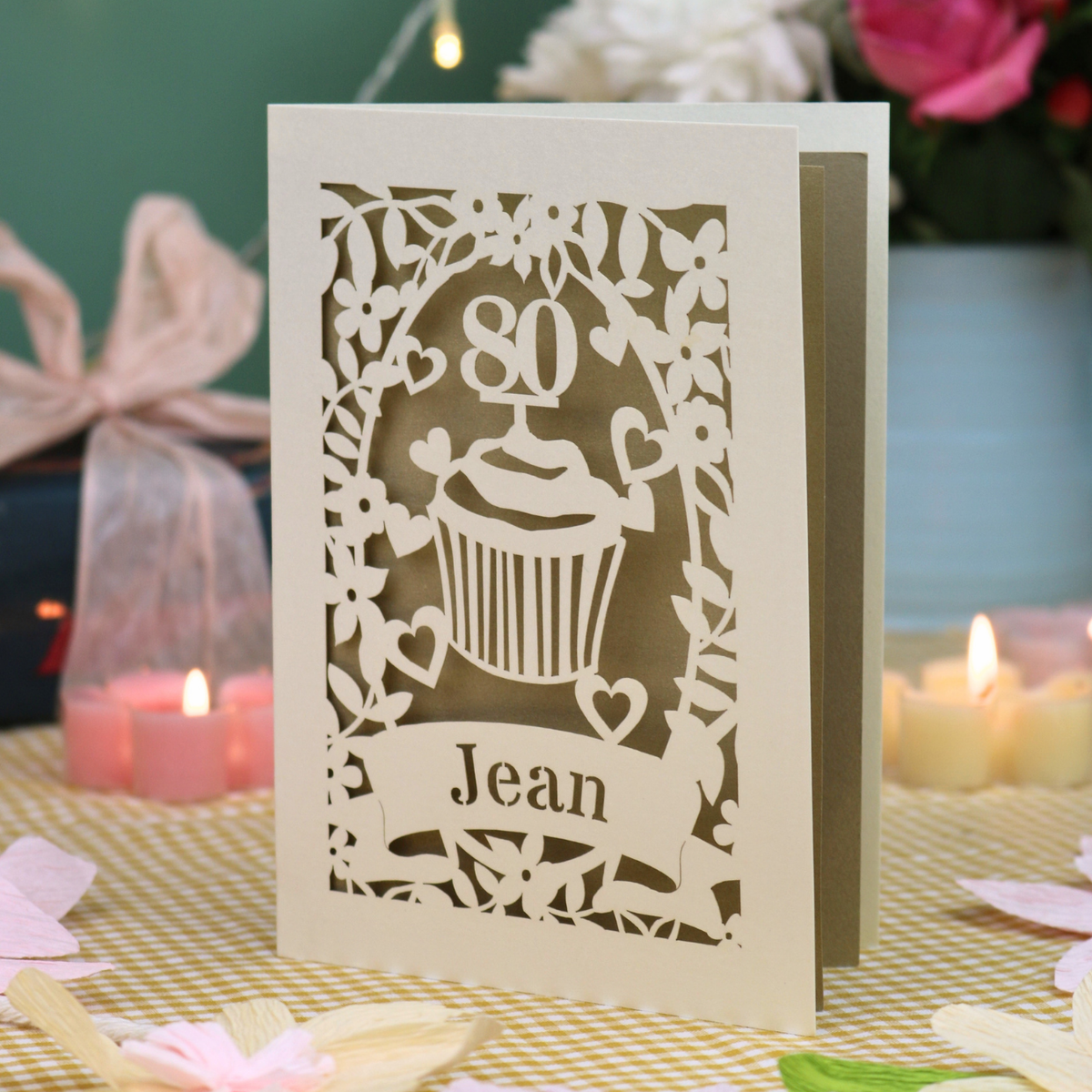 A personalised 80th birthday laser-cut greeting card for Jean, featuring a cream cupcake design with a gold background, displayed on a checked tablecloth with lit candles in the background.