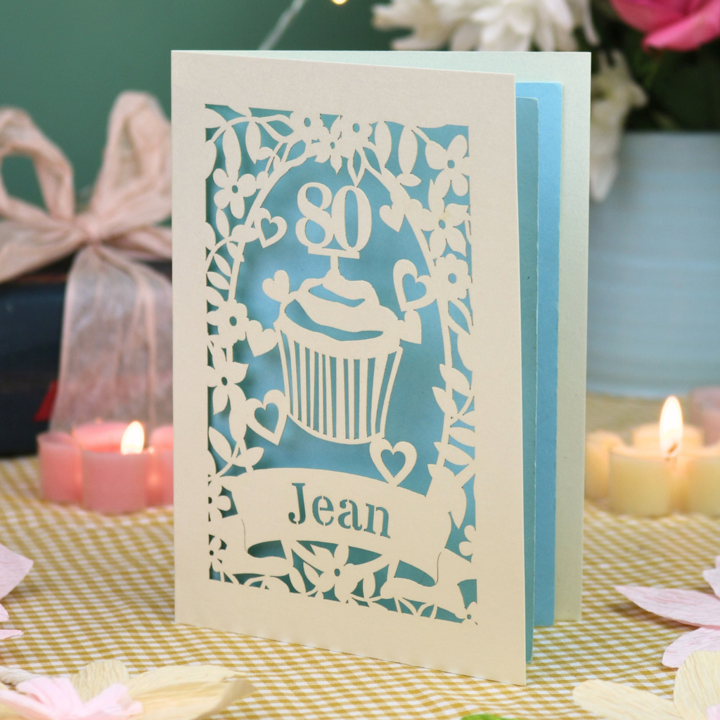 A personalised 80th birthday laser-cut greeting card for Jean, featuring a cream cupcake design with a pastel blue background, displayed on a checked tablecloth with lit candles in the background.