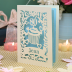 A personalised 80th birthday laser-cut greeting card for Jean, featuring a cream cupcake design with a pastel blue background, displayed on a checked tablecloth with lit candles in the background.