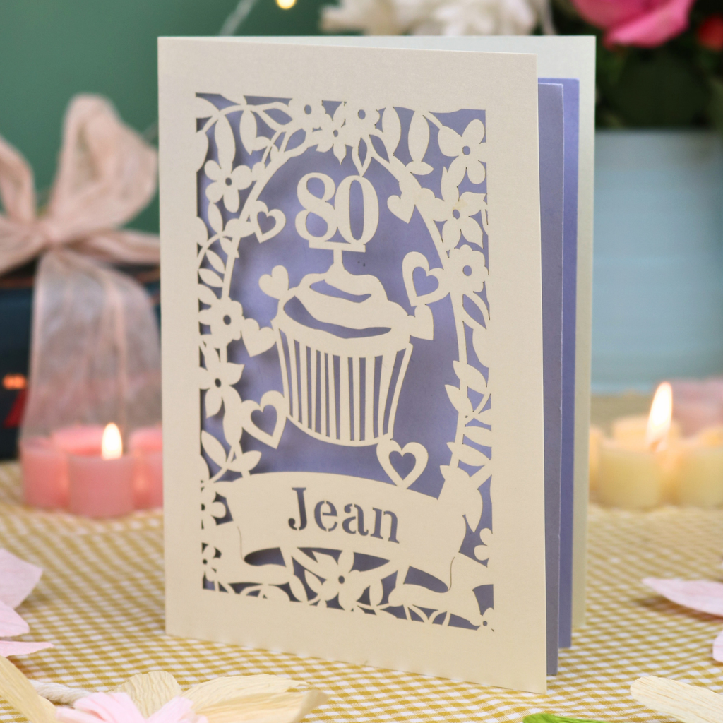 A personalised 80th birthday laser-cut greeting card for Jean, featuring a cream cupcake design with a lilac background, displayed on a checked tablecloth with lit candles in the background.