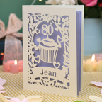 A personalised 80th birthday laser-cut greeting card for Jean, featuring a cream cupcake design with a lilac background, displayed on a checked tablecloth with lit candles in the background.