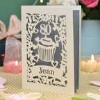 A personalised 80th birthday laser-cut greeting card for Jean, featuring a cream cupcake design with a silver background, displayed on a checked tablecloth with lit candles in the background.