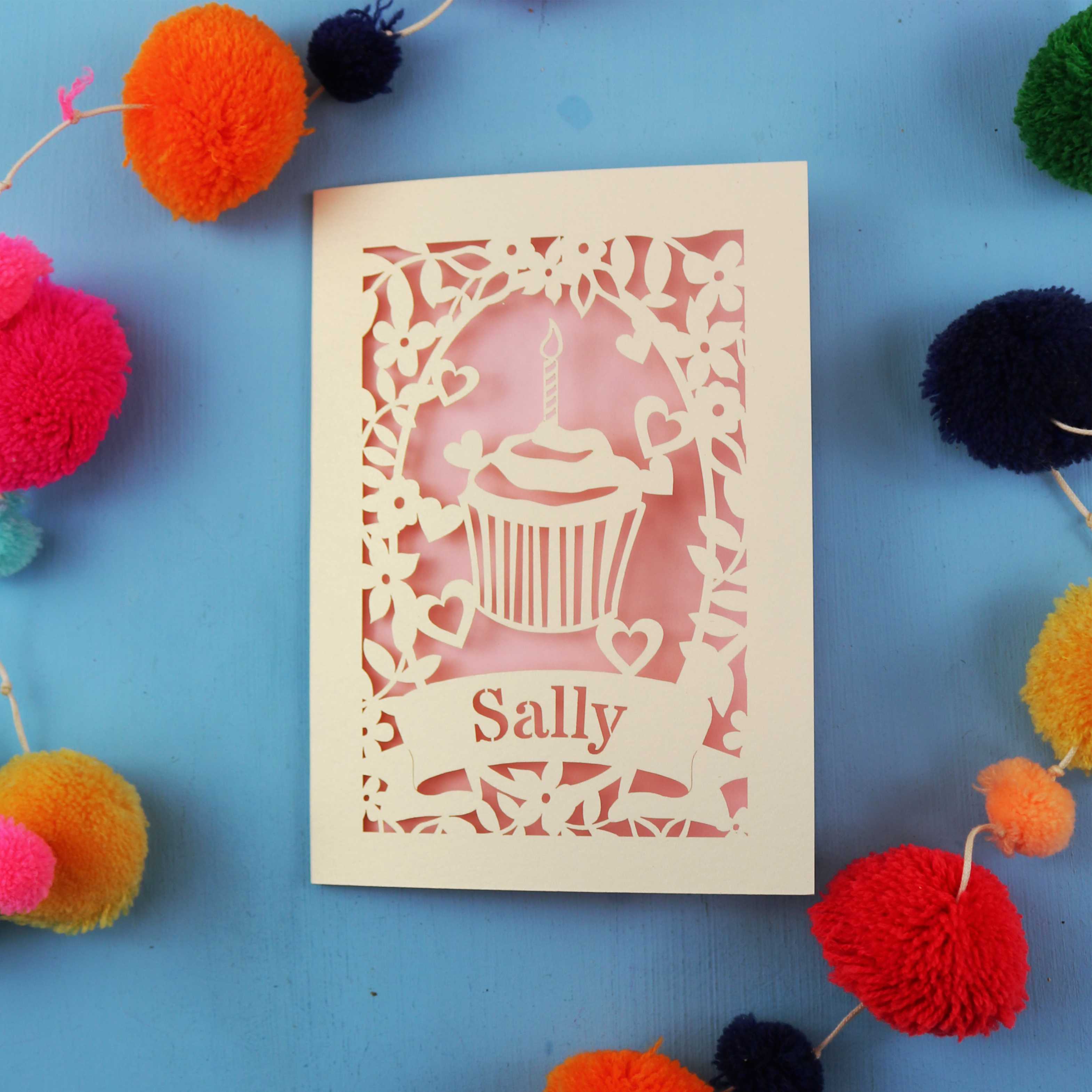 Laser Cut birthday card with a cupcake design and 'Sally' text, surrounded by colourful pom-poms on a blue background.
