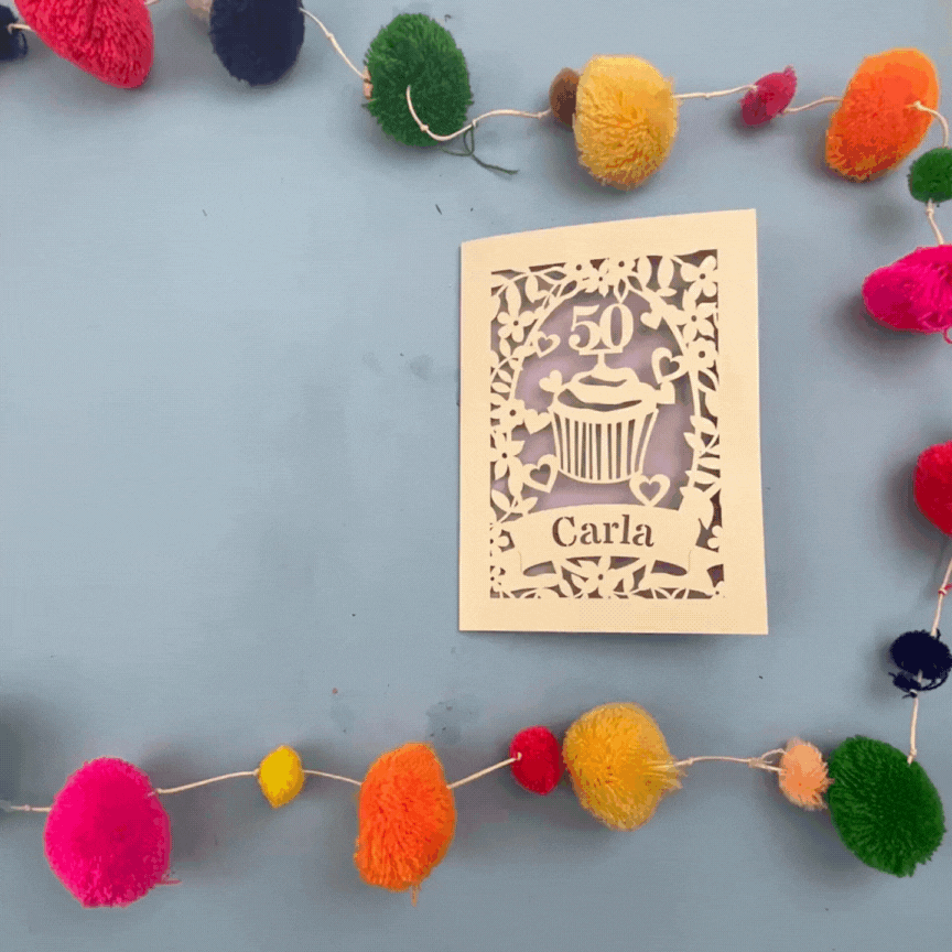 Decorative laser cut birthday card with '50' and 'Carla' on a blue background with colourful pom-poms.
