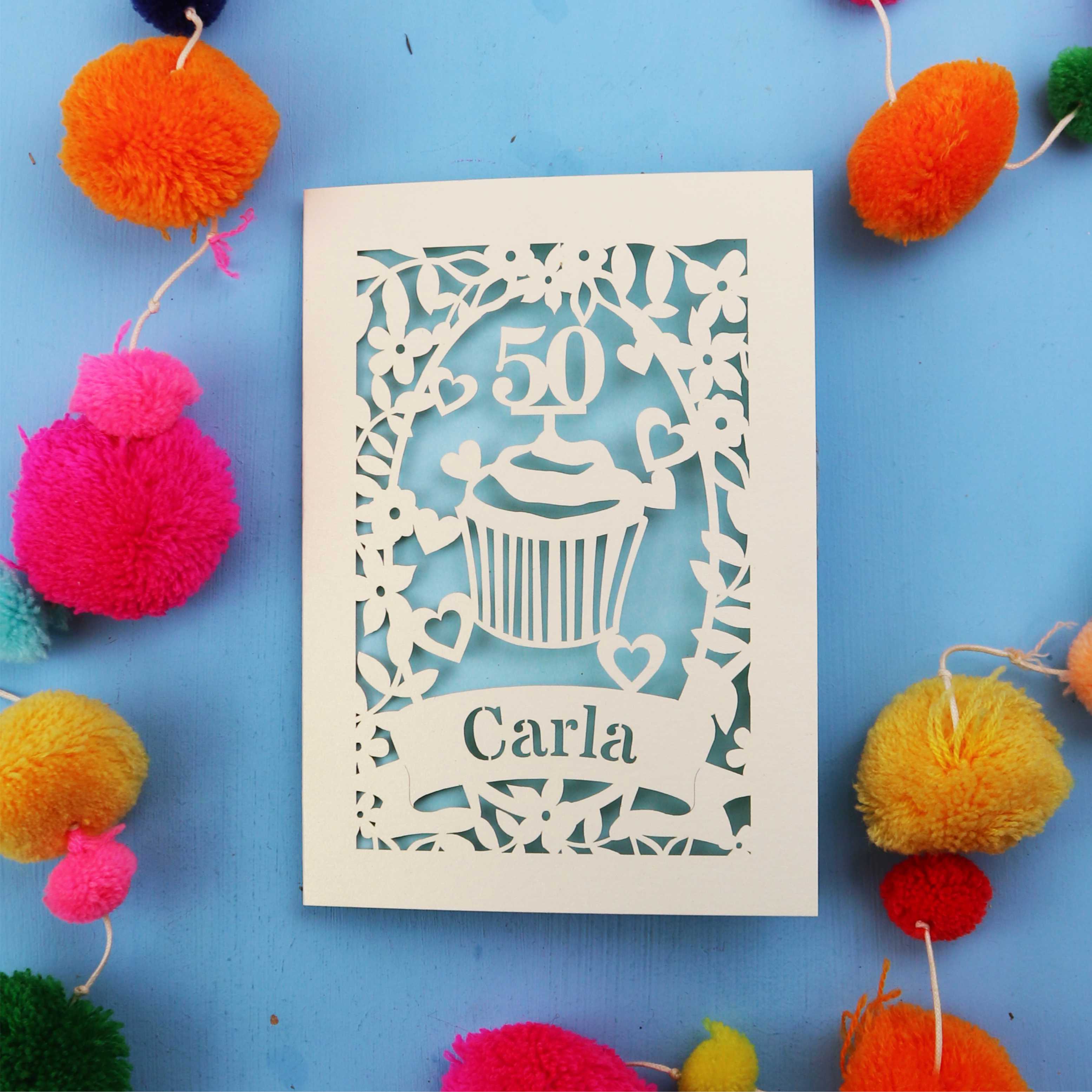 Decorative birthday card with '50' and 'Carla' on a blue background with colourful pom-poms.