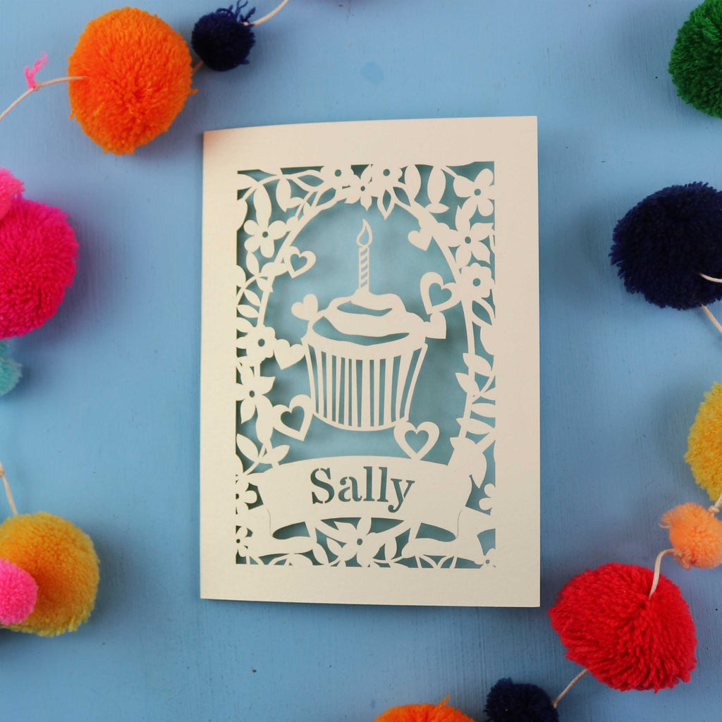 Unique birthday card with a cupcake design and 'Sally' text, surrounded by colourful pom-poms on a blue background.