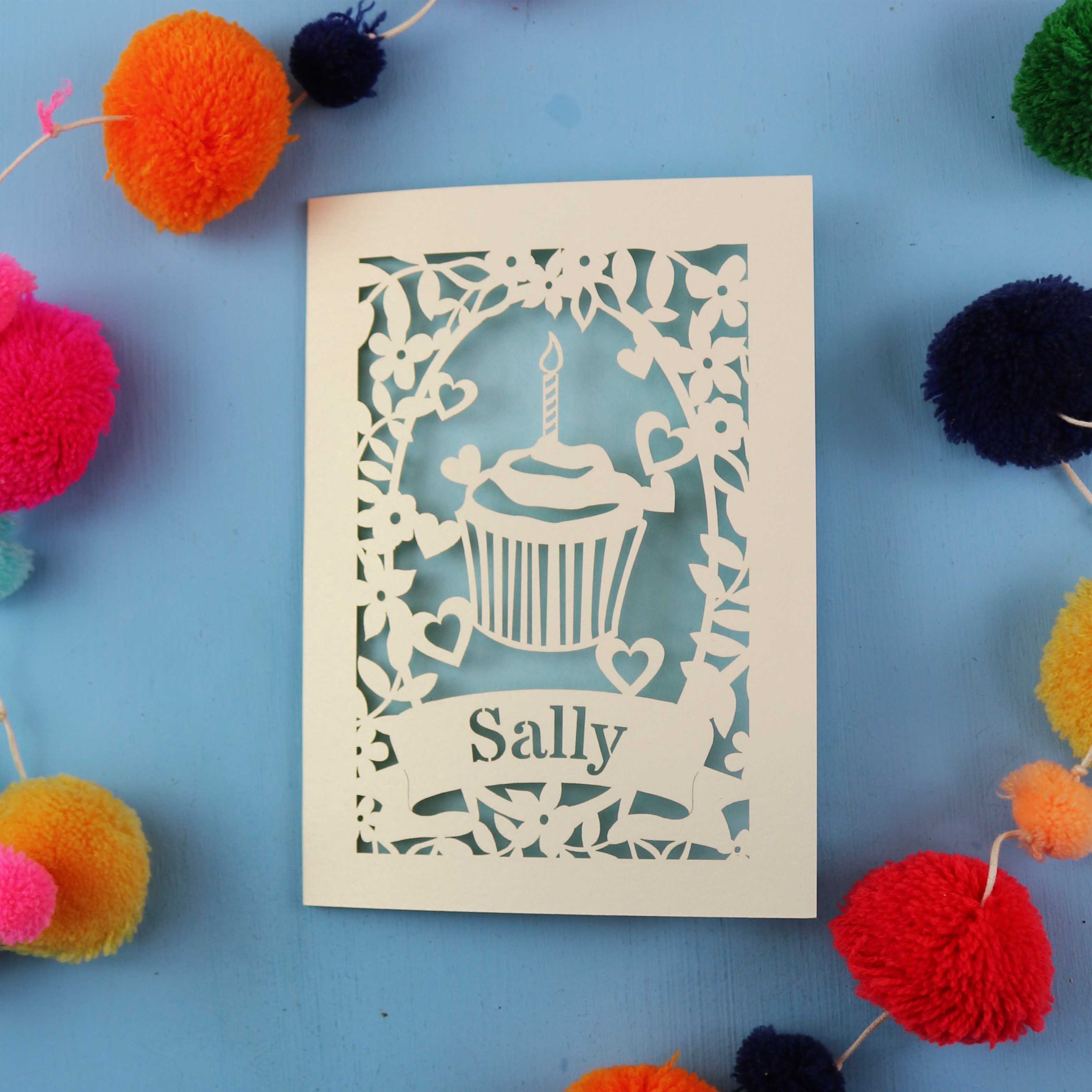 Unique birthday card with a cupcake design and 'Sally' text, surrounded by colourful pom-poms on a blue background.
