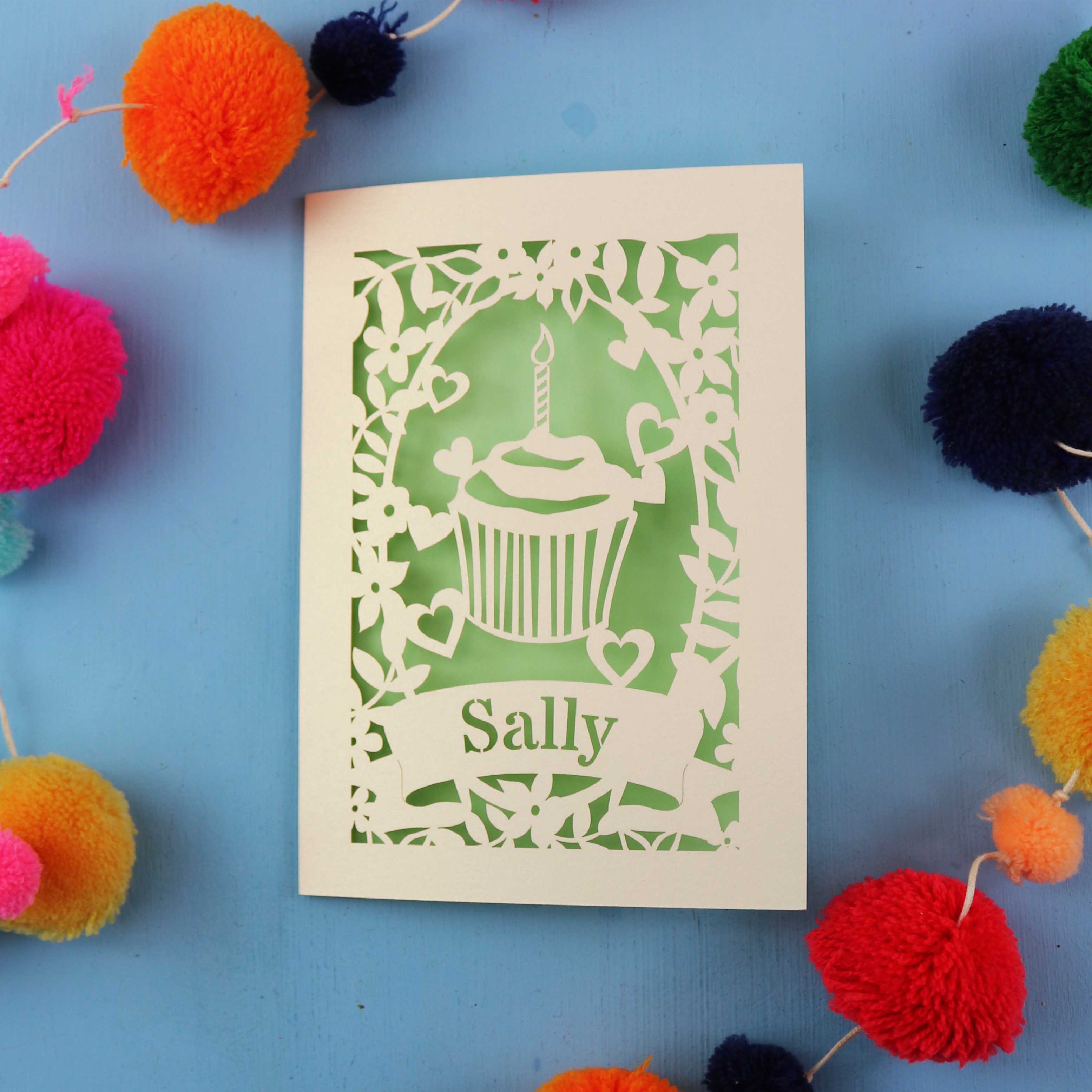 Personalised Cupcake birthday card with a cupcake design and 'Sally' text, surrounded by colourful pom-poms on a blue background.