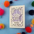 Personalised birthday card with a cupcake design and 'Sally' text, surrounded by colourful pom-poms on a blue background.