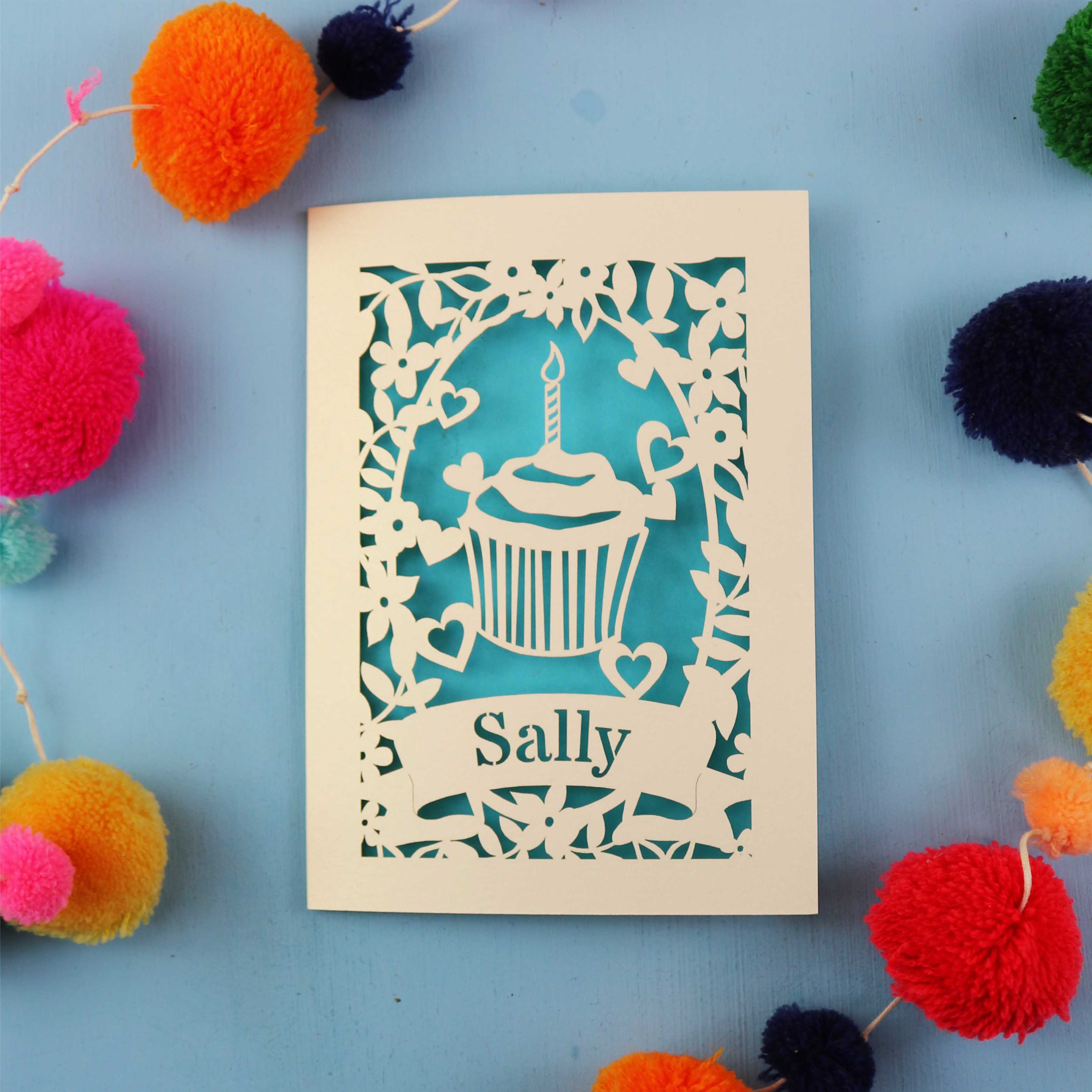 Cute birthday card with a cupcake design and 'Sally' text, surrounded by colourful pom-poms on a blue background.