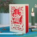 A laser cut birthday card with floral border, cupcake design and personalised with the name "Fiona" cut out of a banner shape