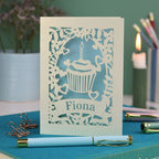 A laser cut birthday card with floral border, cupcake design and personalised with the name "Fiona" cut out of a banner shape