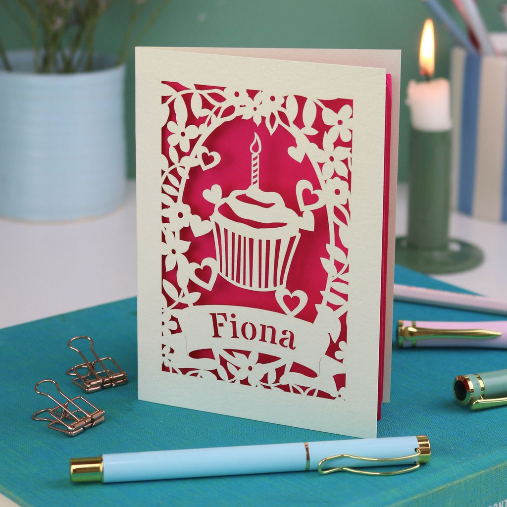 A laser cut birthday card with floral border, cupcake design and personalised with the name "Fiona" cut out of a banner shape