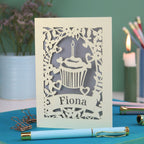 A laser cut birthday card with floral border, cupcake design and personalised with the name "Fiona" cut out of a banner shape
