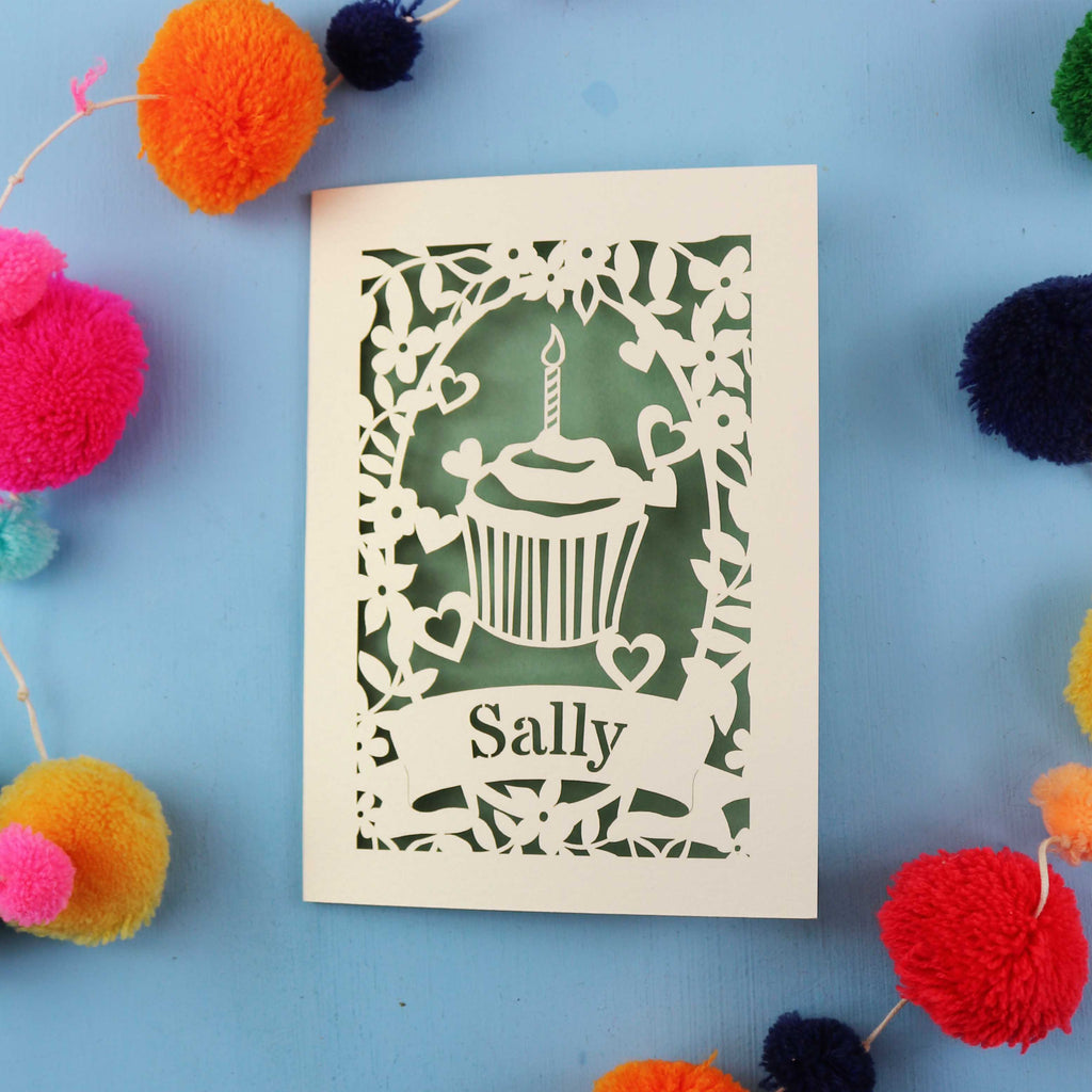 Unique decorative birthday card with a cupcake design and 'Sally' text, surrounded by colourful pom-poms on a blue background.