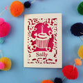 Decorative birthday card with a cupcake design and 'Sally' text, surrounded by colourful pom-poms on a blue background.