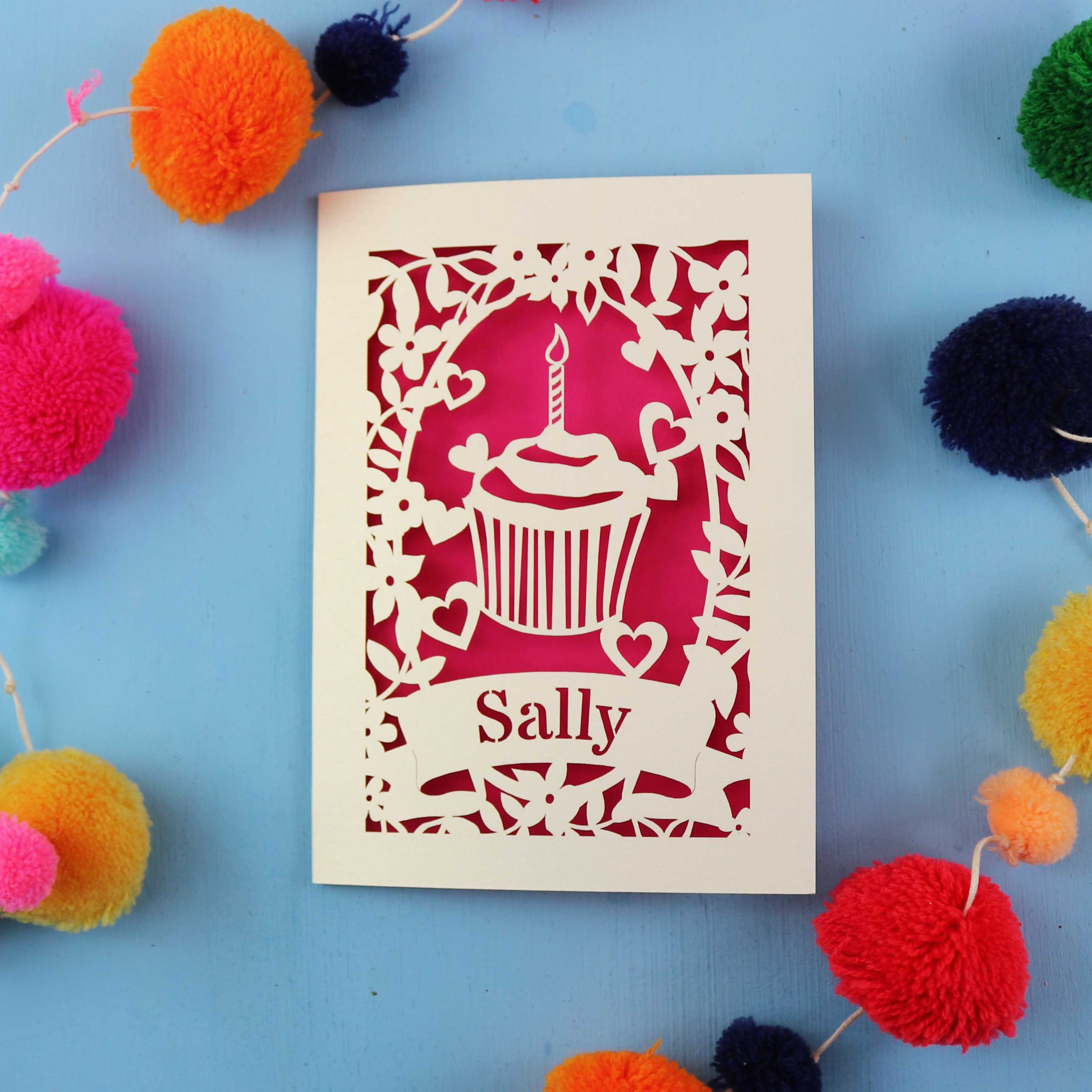 Decorative birthday card with a cupcake design and 'Sally' text, surrounded by colourful pom-poms on a blue background.