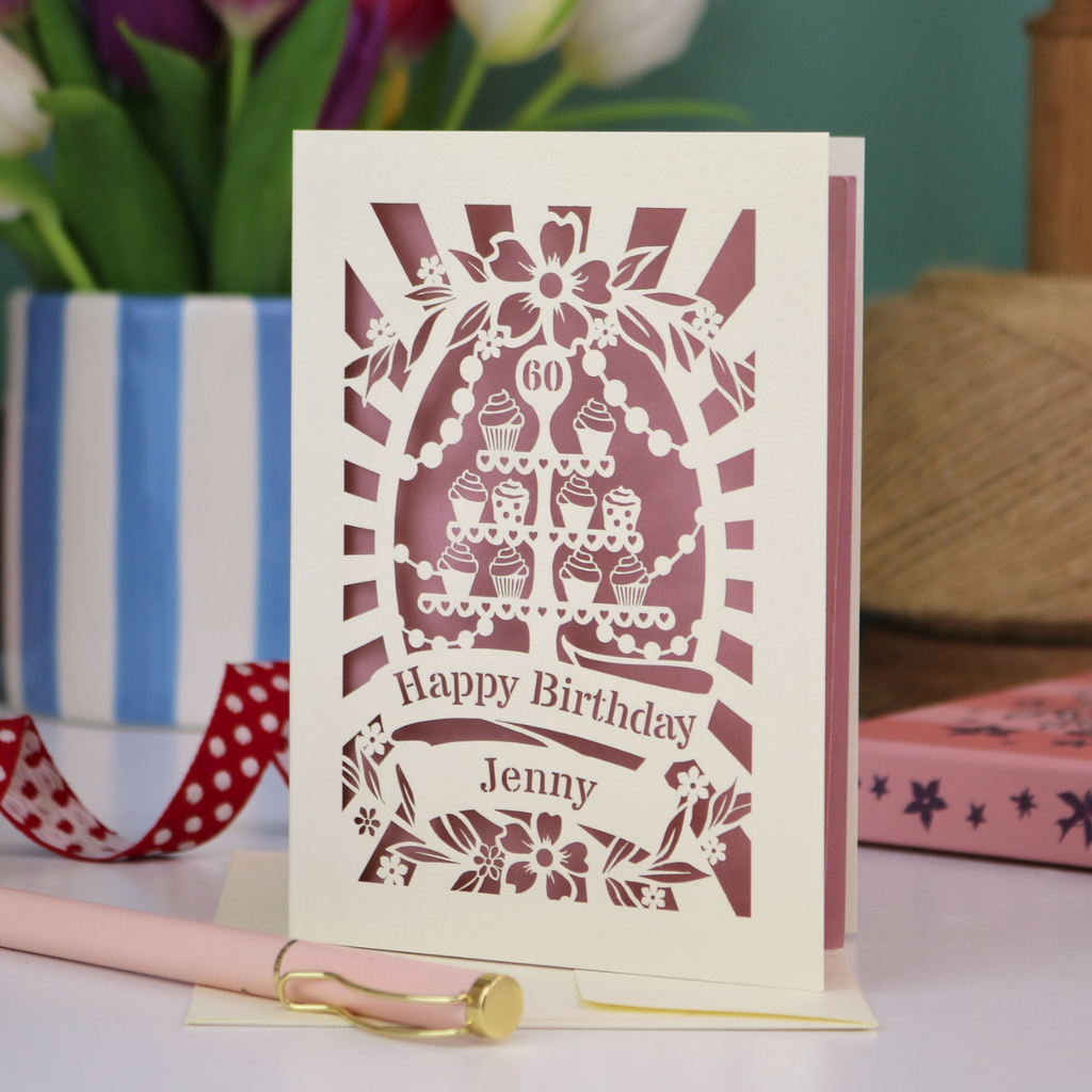 A cream and dusky pink laser cut 60th birthday card with cupcakes on a cake stand design, the number '60' and "Happy Birthday Jenny" personalised text in cut out stencil lettering