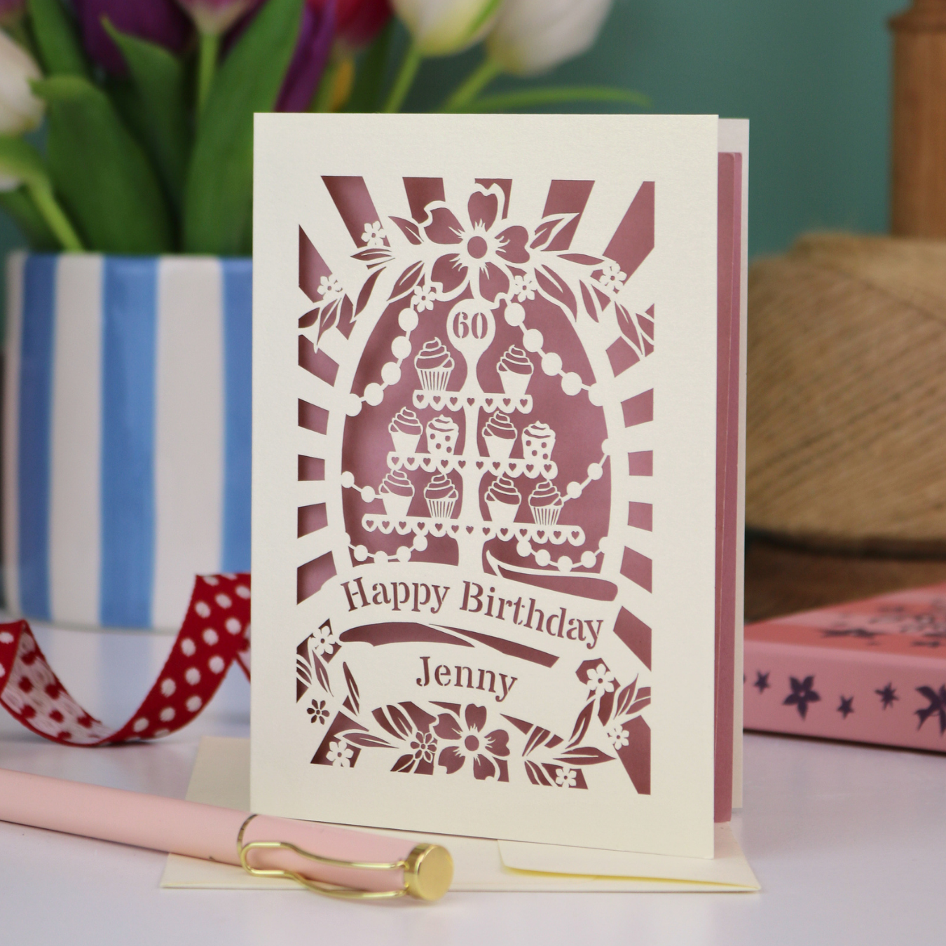 A cream and dusky pink laser cut 60th birthday card with cupcakes on a cake stand design, the number '60' and "Happy Birthday Jenny" personalised text in cut out stencil lettering