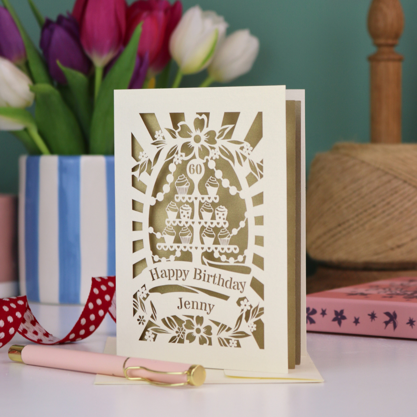 A cream and gold leaf laser cut 60th birthday card with cupcakes on a cake stand design, the number '60' and "Happy Birthday Jenny" personalised text in cut out stencil lettering