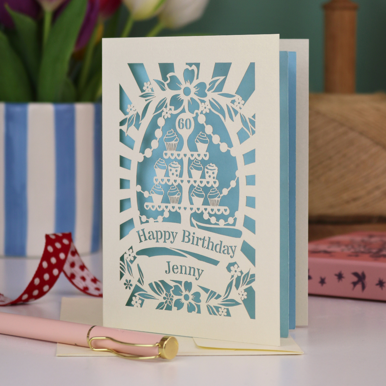 A cream and light blue laser cut 60th birthday card with cupcakes on a cake stand design, the number '60' and "Happy Birthday Jenny" personalised text in cut out stencil lettering