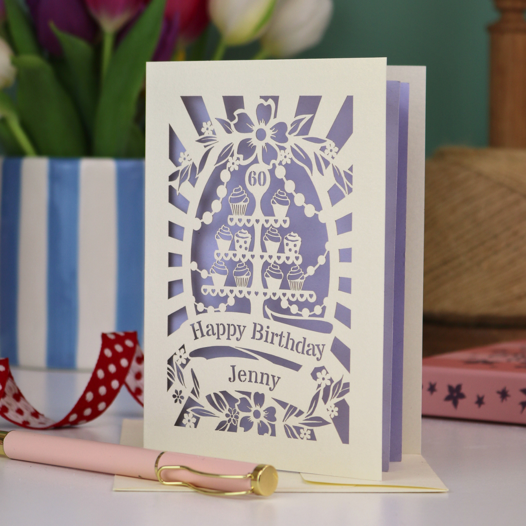 A cream and lilac laser cut personalised 60th birthday card with cupcakes on a cake stand design, the number '60' and "Happy Birthday Jenny" personalised text in cut out stencil lettering