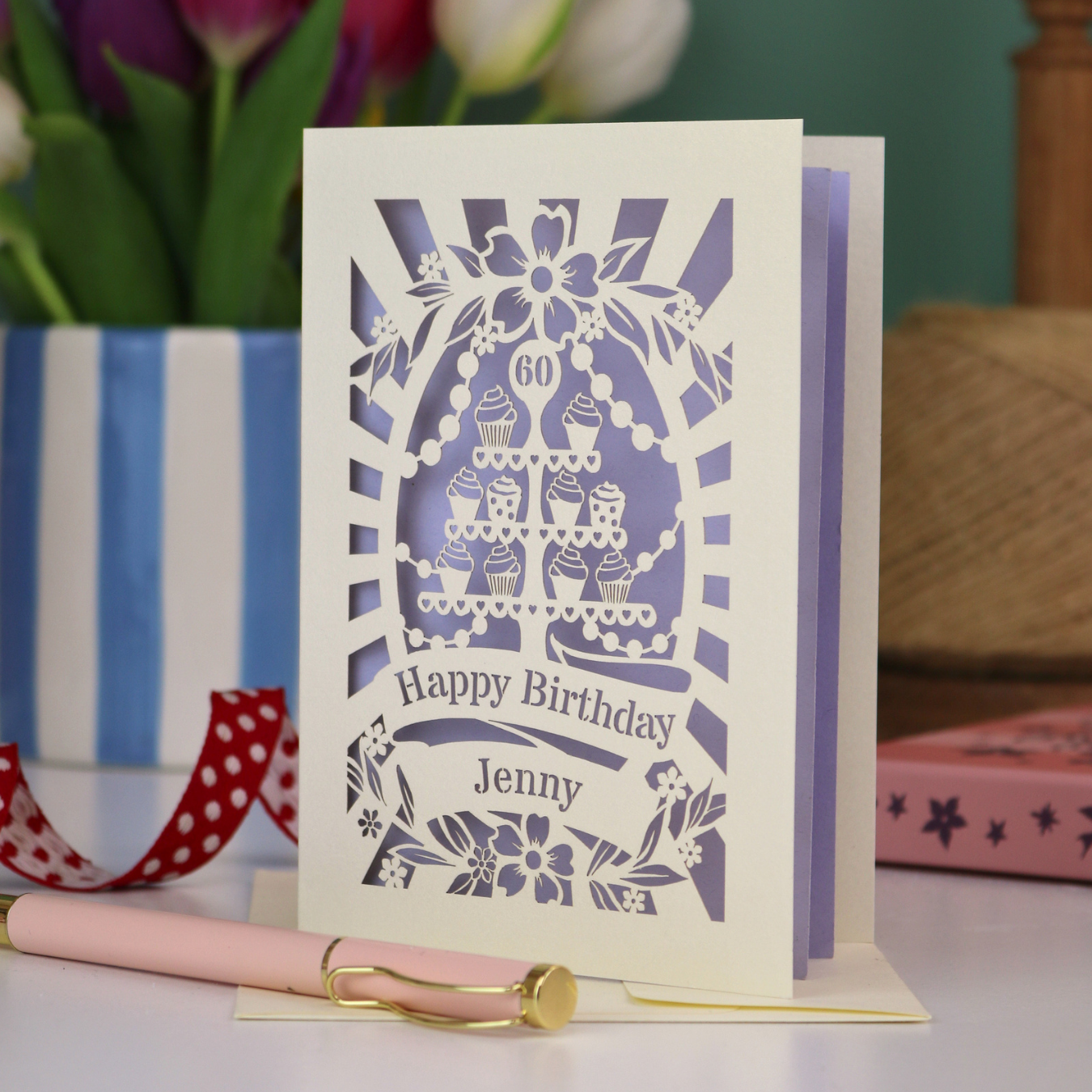 A cream and lilac laser cut personalised 60th birthday card with cupcakes on a cake stand design, the number '60' and "Happy Birthday Jenny" personalised text in cut out stencil lettering