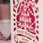 A cream and shocking pink laser cut 60th birthday card with cupcakes on a cake stand design, the number '60' and "Happy Birthday Jenny" personalised text in cut out stencil lettering