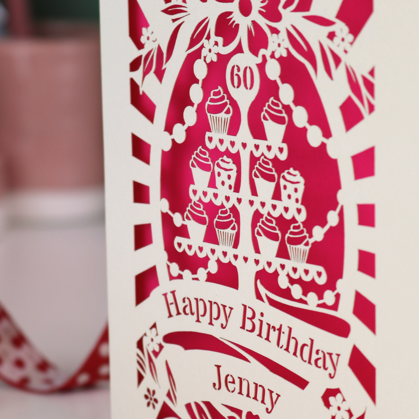 A cream and shocking pink laser cut 60th birthday card with cupcakes on a cake stand design, the number '60' and "Happy Birthday Jenny" personalised text in cut out stencil lettering