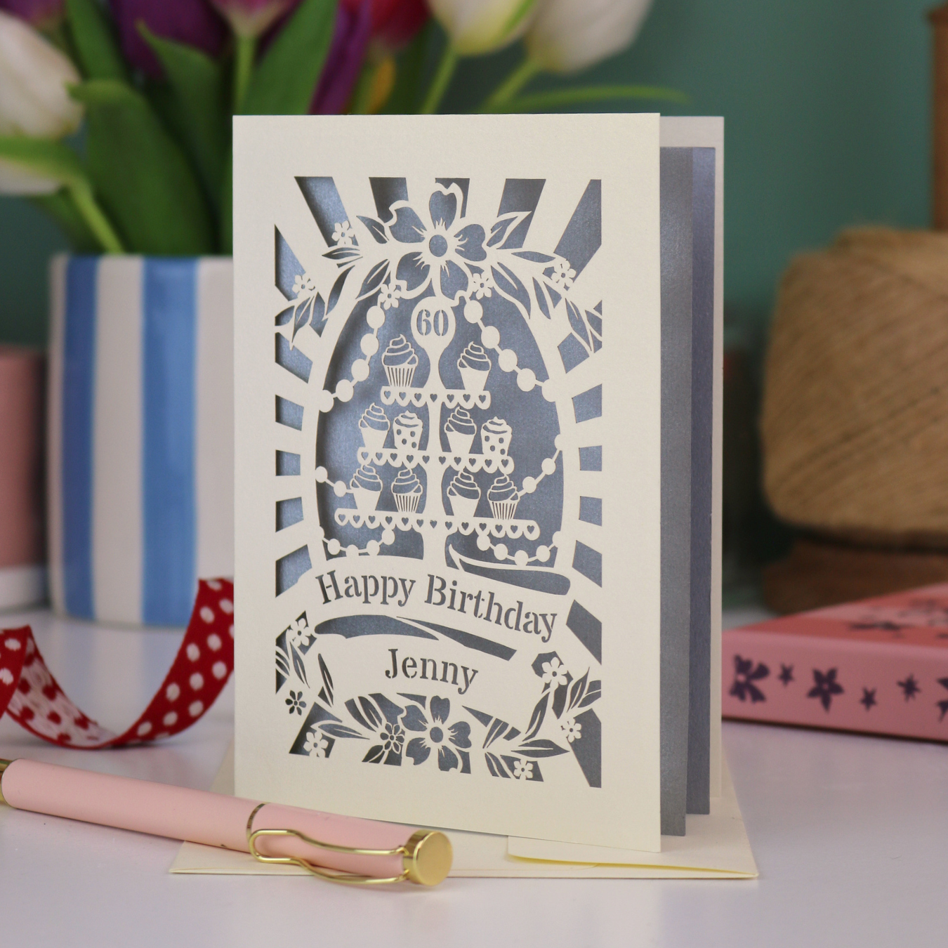A cream and silver laser cut 60th birthday card with cupcakes on a cake stand design, the number '60' and "Happy Birthday Jenny" personalised text in cut out stencil lettering
