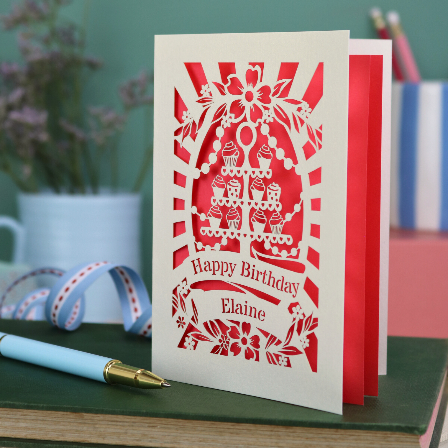 Laser cut Birthday card with cream cupcake design and text 'Happy Birthday Elaine' on a green surface with a pen and flowers.