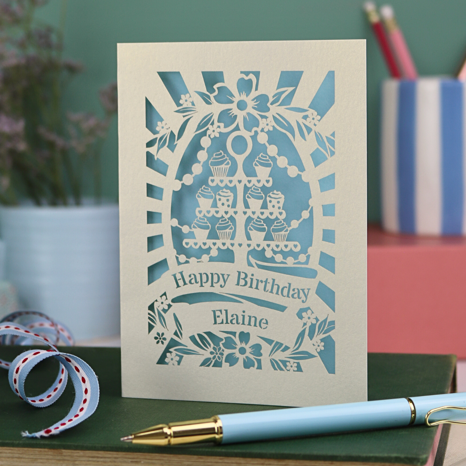 Personalised birthday card with blue cut-out design on a green surface with a pen and decorative ribbon.