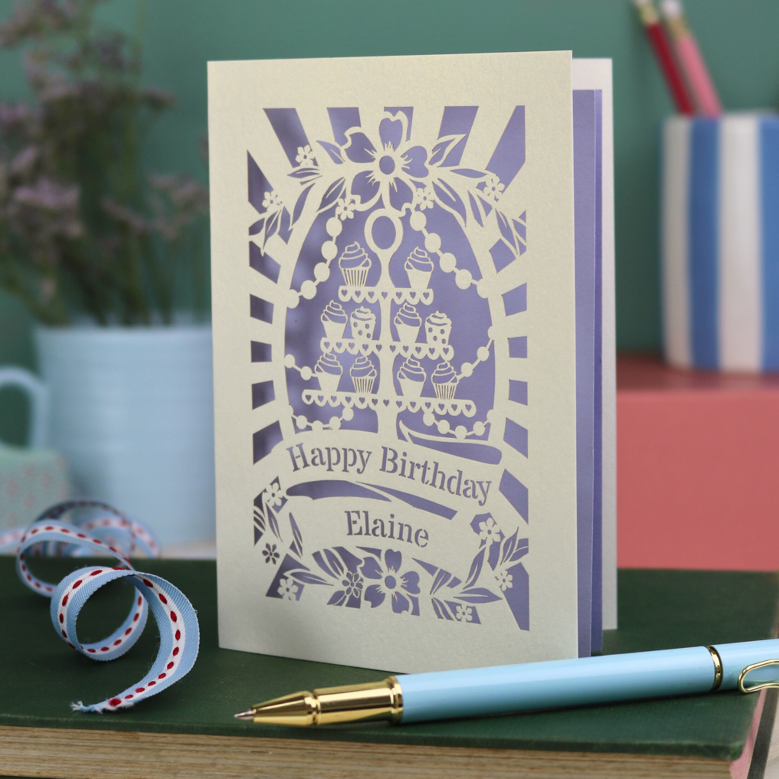 Laser cut Birthday card with cream cupcake design and text 'Happy Birthday Elaine' on a green surface with a pen and flowers.