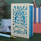 Laser-cut cupcake birthday card with blue design on a green surface with stationery items.