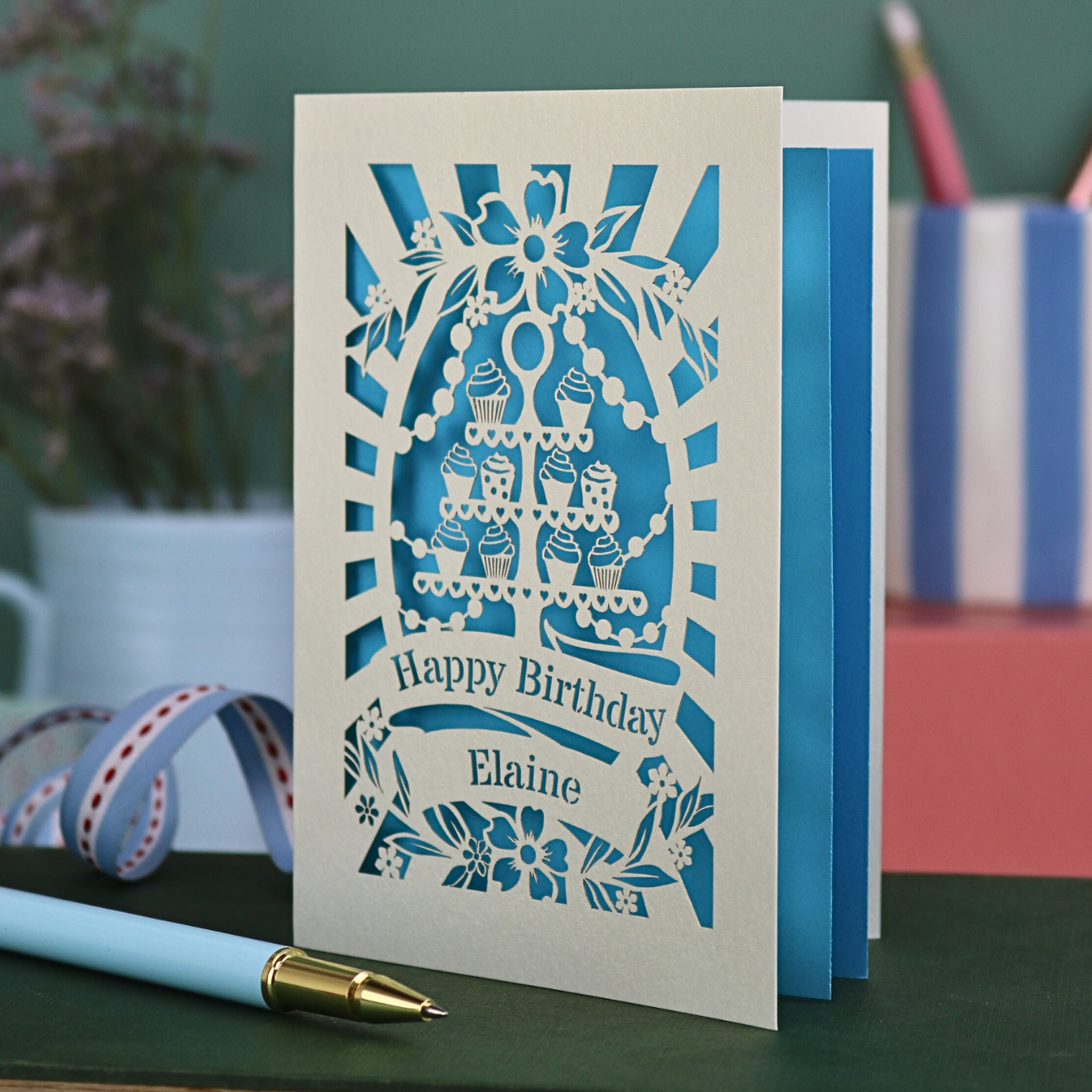 Laser-cut cupcake birthday card with blue design on a green surface with stationery items.