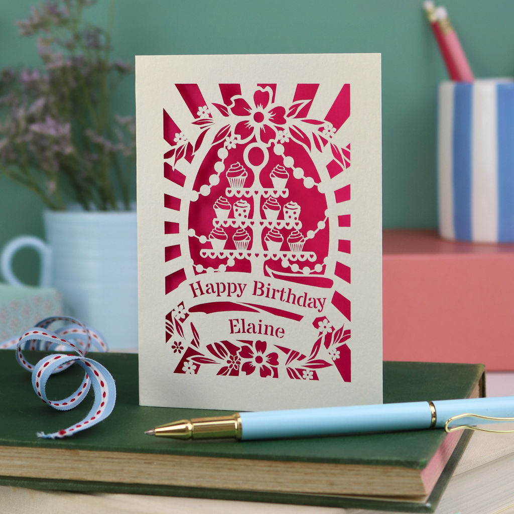 Laser cut Birthday card with cream cupcake design and text 'Happy Birthday Elaine' on a green surface with a pen and flowers.