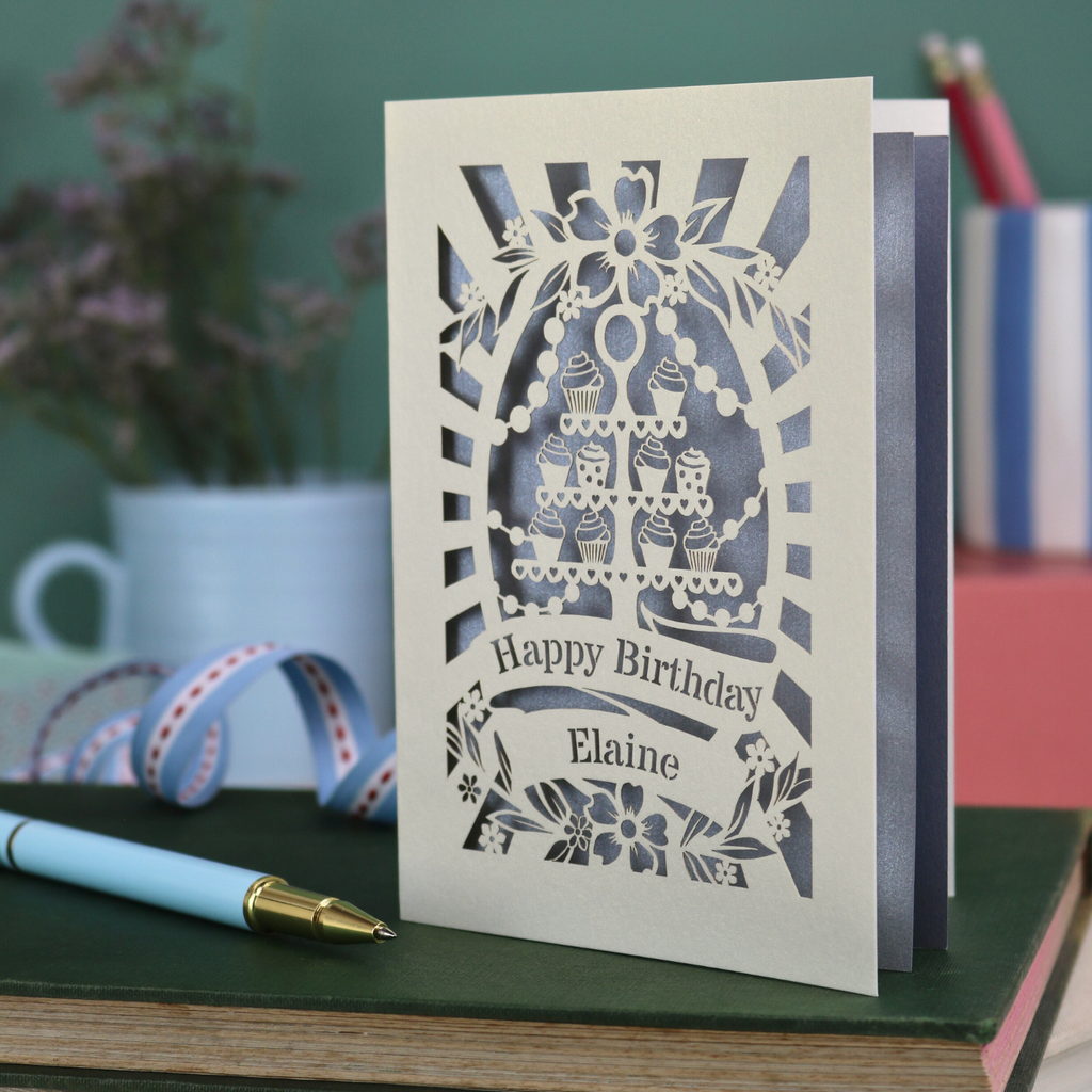 Laser cut Birthday card with cream cupcake design and text 'Happy Birthday Elaine' on a green surface with a pen and flowers.
