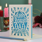 A cream Christmas greeting card with intricate laser-cut snowflake and floral patterns, featuring the text 'MERRY Christmas to MY DAD' over a bright blue background. The card is displayed on a tabletop with a pencil in the foreground and blurred holiday decorations, including candles and a red ribbon, in the background.