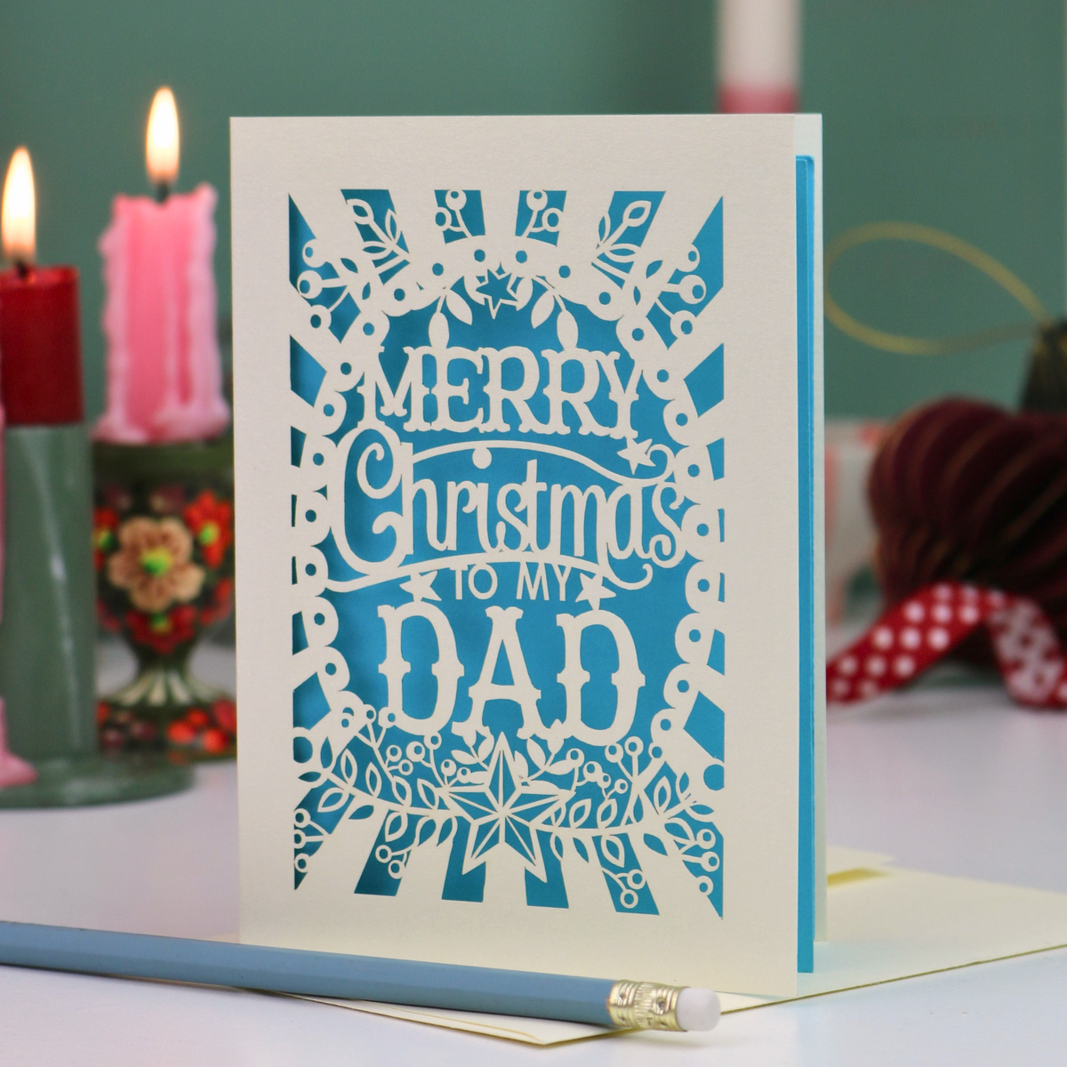 A cream Christmas greeting card with intricate laser-cut snowflake and floral patterns, featuring the text 'MERRY Christmas to MY DAD' over a bright blue background. The card is displayed on a tabletop with a pencil in the foreground and blurred holiday decorations, including candles and a red ribbon, in the background.