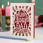 A laser-cut Christmas greeting card with the message 'MERRY Christmas TO MY DAD' in white cutout lettering over a deep red background, displayed on a tabletop with candles and a pencil in a softly blurred holiday setting.
