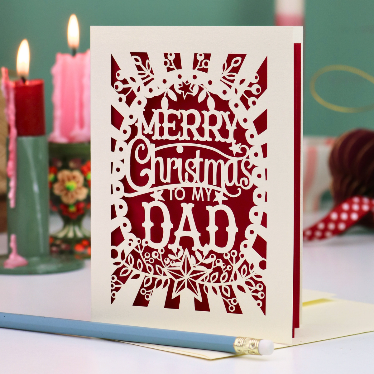 A laser-cut Christmas greeting card with the message 'MERRY Christmas TO MY DAD' in white cutout lettering over a deep red background, displayed on a tabletop with candles and a pencil in a softly blurred holiday setting.
