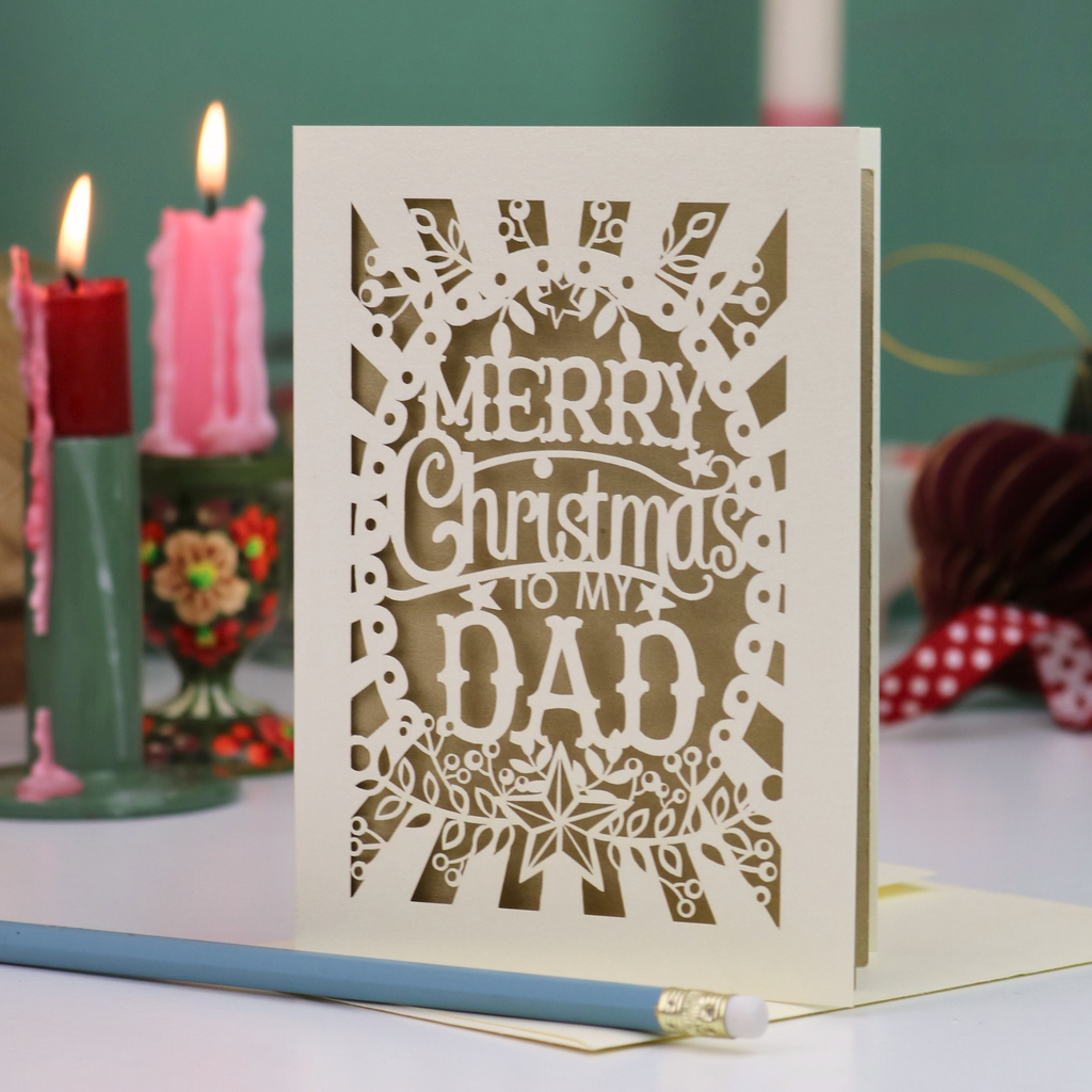 A laser-cut Christmas greeting card that reads 'MERRY Christmas TO MY DAD' in white cutout lettering on an off-white card, standing on a table with candles and holiday decor blurred in the background.