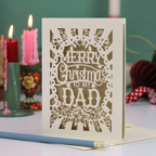 A laser-cut Christmas greeting card that reads 'MERRY Christmas TO MY DAD' in white cutout lettering on an off-white card, standing on a table with candles and holiday decor blurred in the background.