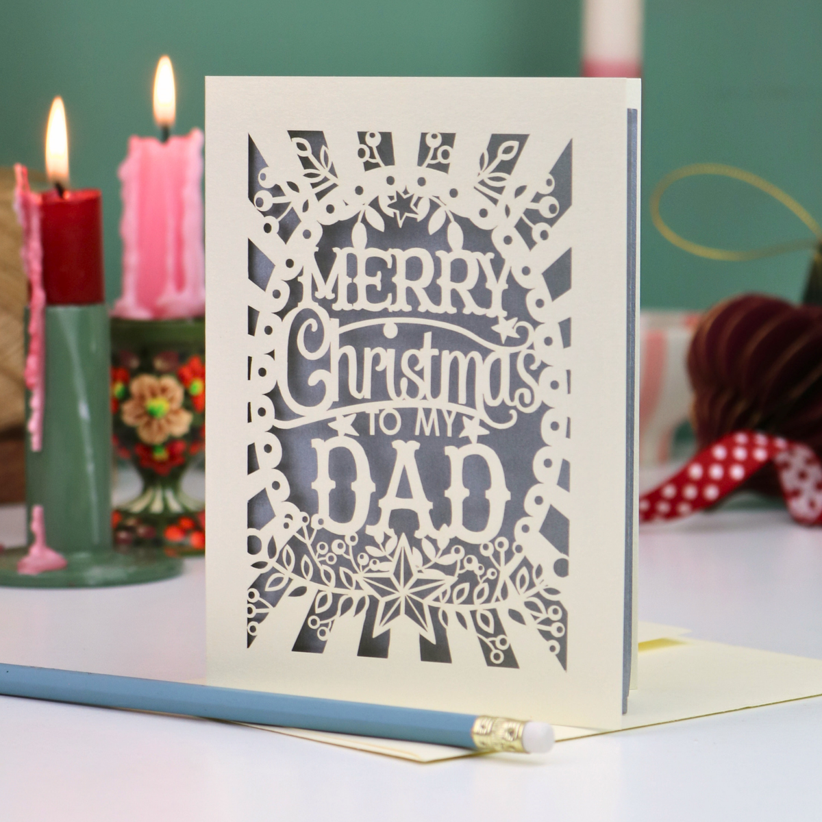 A laser-cut Christmas greeting card with intricate cutout patterns and the text 'MERRY Christmas TO MY DAD' in white, displayed on a tabletop with candles and stationery in the background.
