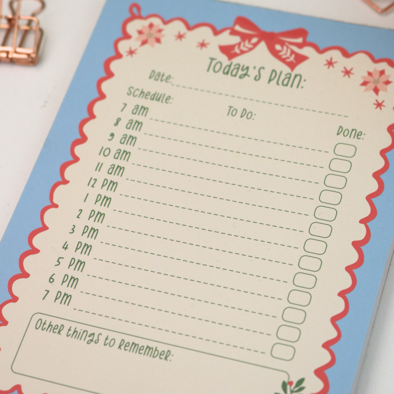 A close-up of a decorative daily planner notepad with a light blue cover and a cream page featuring a red scalloped border, floral accents, and time slots for scheduling and reminders.