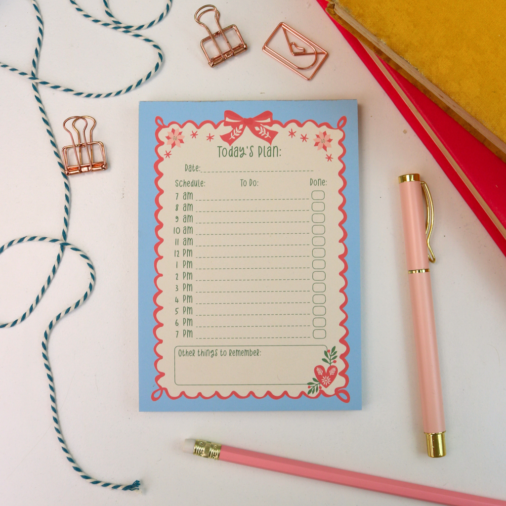 A flat-lay view of a 'Today's Plan' notepad with a light blue background and pink/red decorative border, styled on a white surface with a pink pen, binder clips, twine, and books.