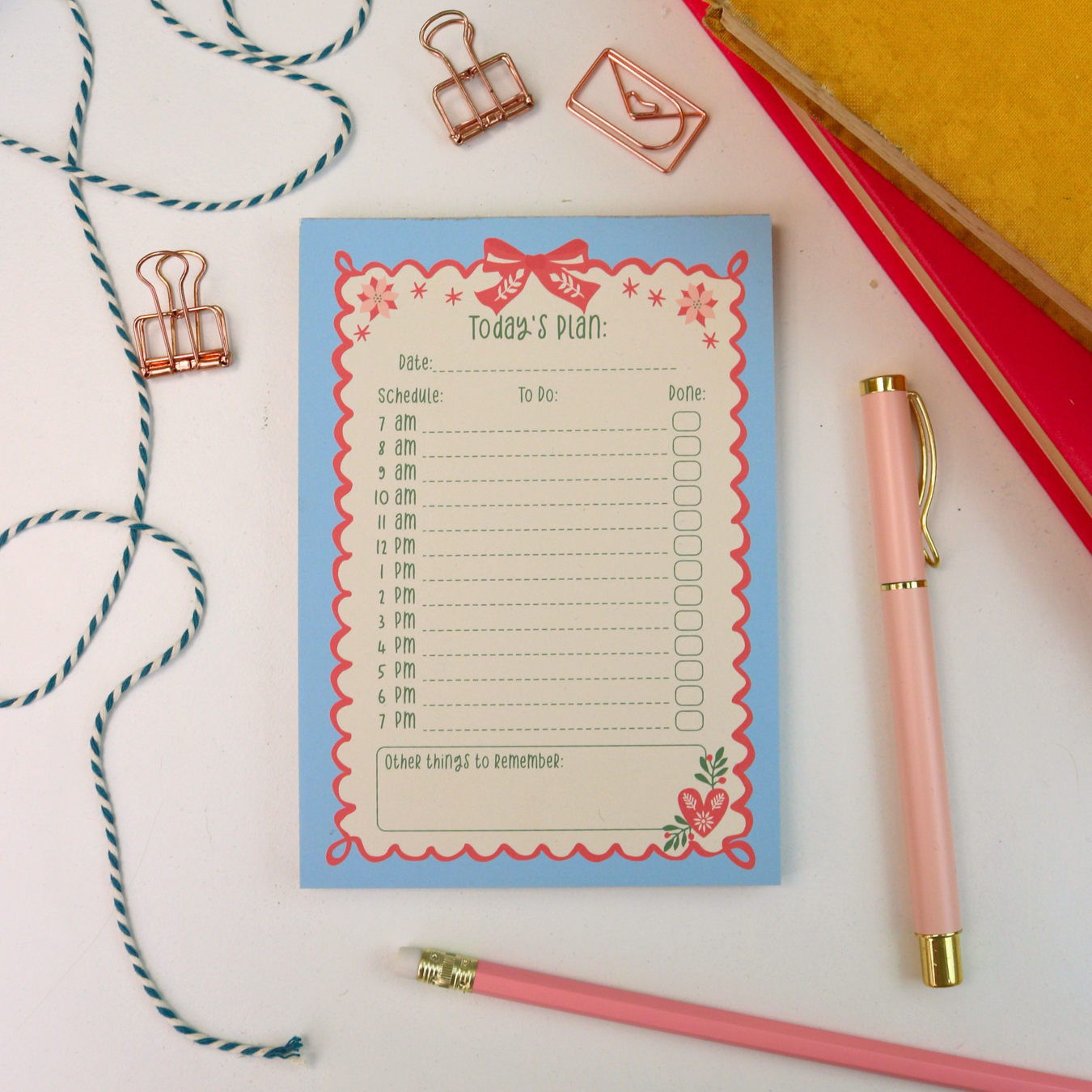 A flat-lay view of a 'Today's Plan' notepad with a light blue background and pink/red decorative border, styled on a white surface with a pink pen, binder clips, twine, and books.