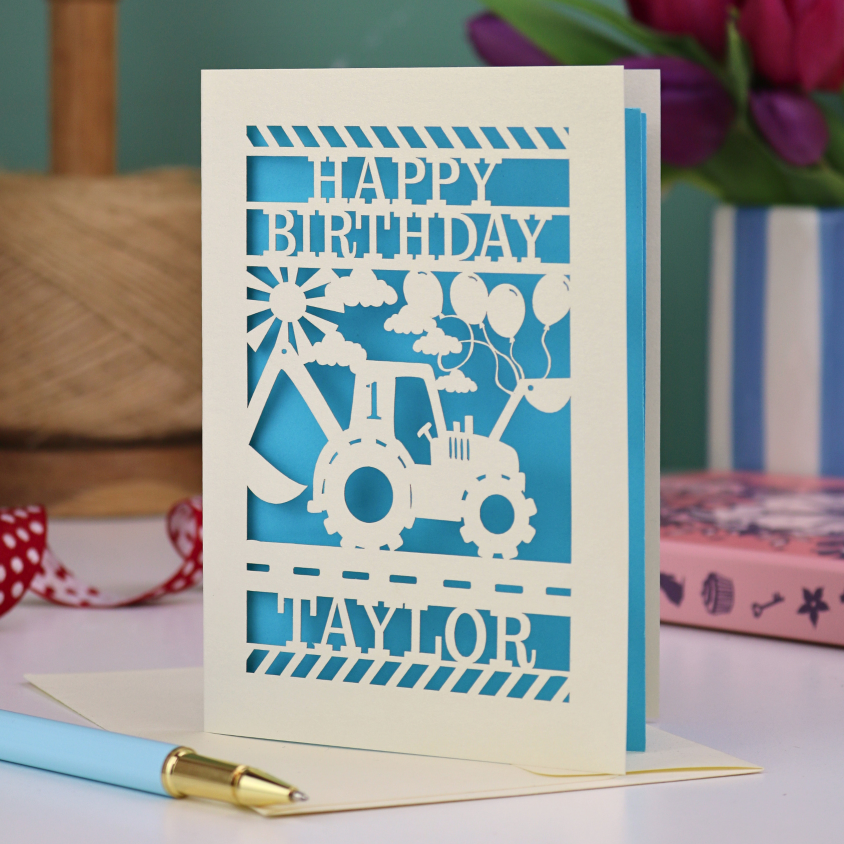 1st Birthday card with tractor design and 'Happy Birthday Taylor' text on a table with a pen and books.